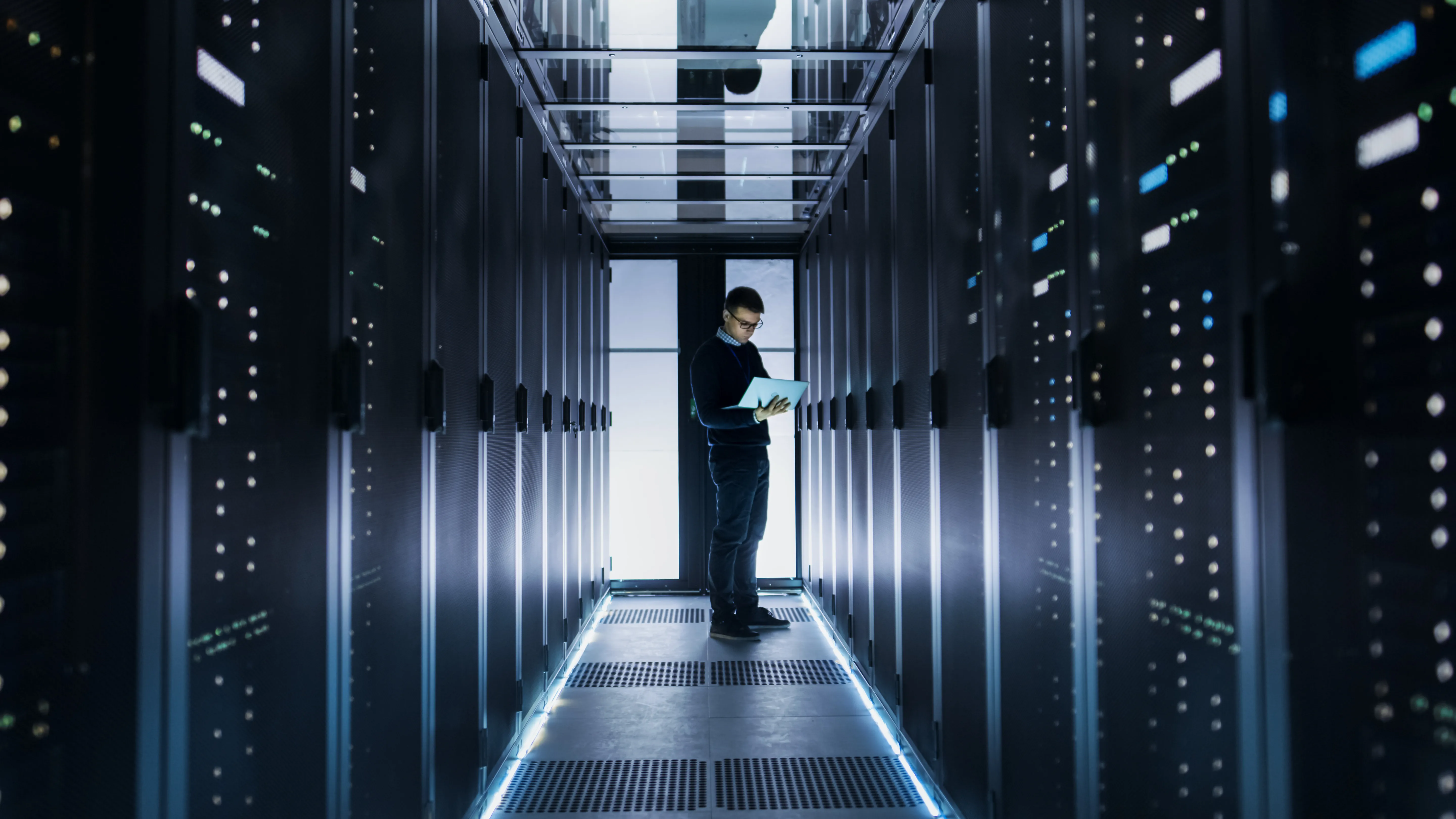 Male IT engineer works on a laptop at the end of a corridor in a big data center. Rows of rack servers are seen.